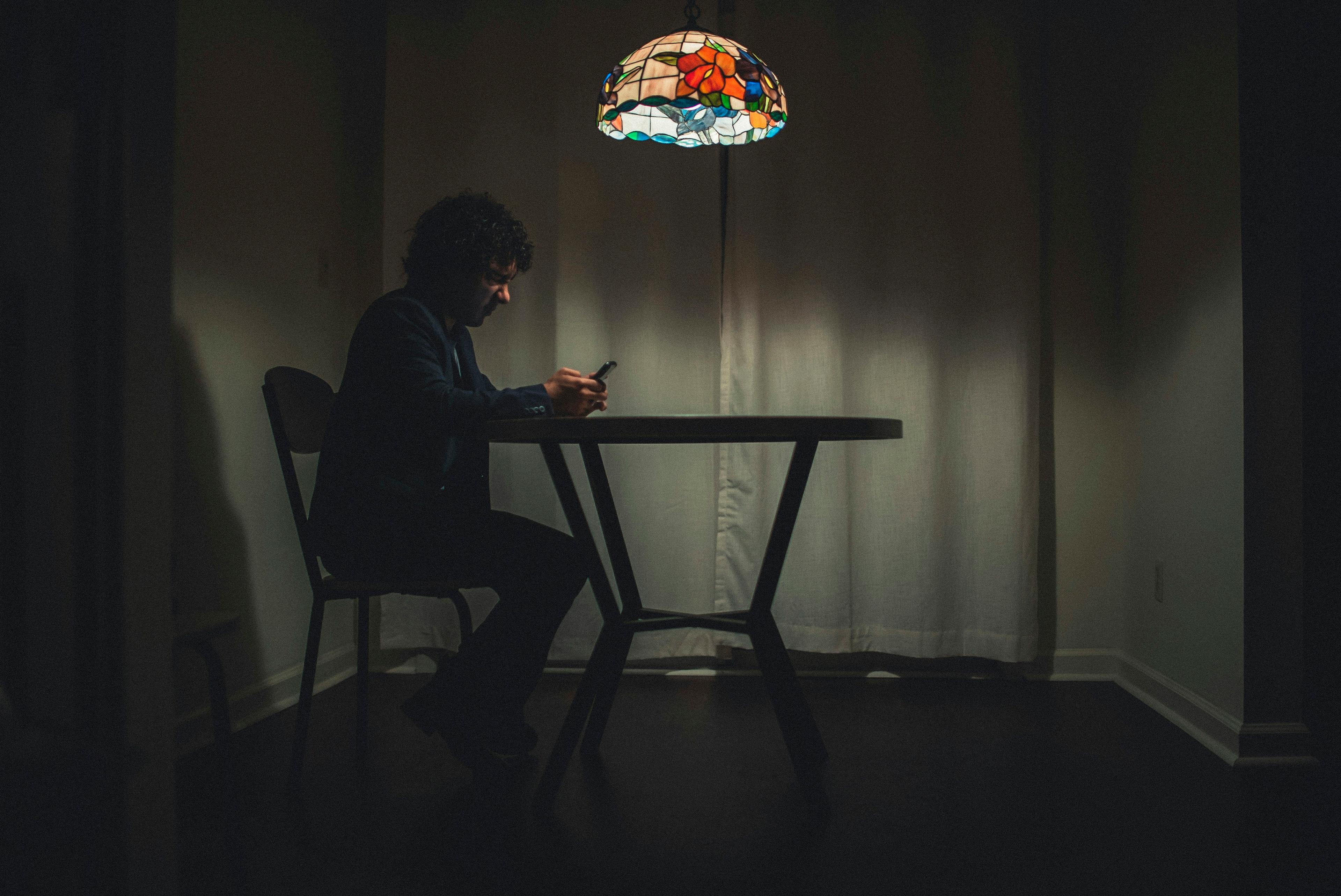 A man sits alone at a table in a dark room under a lamp